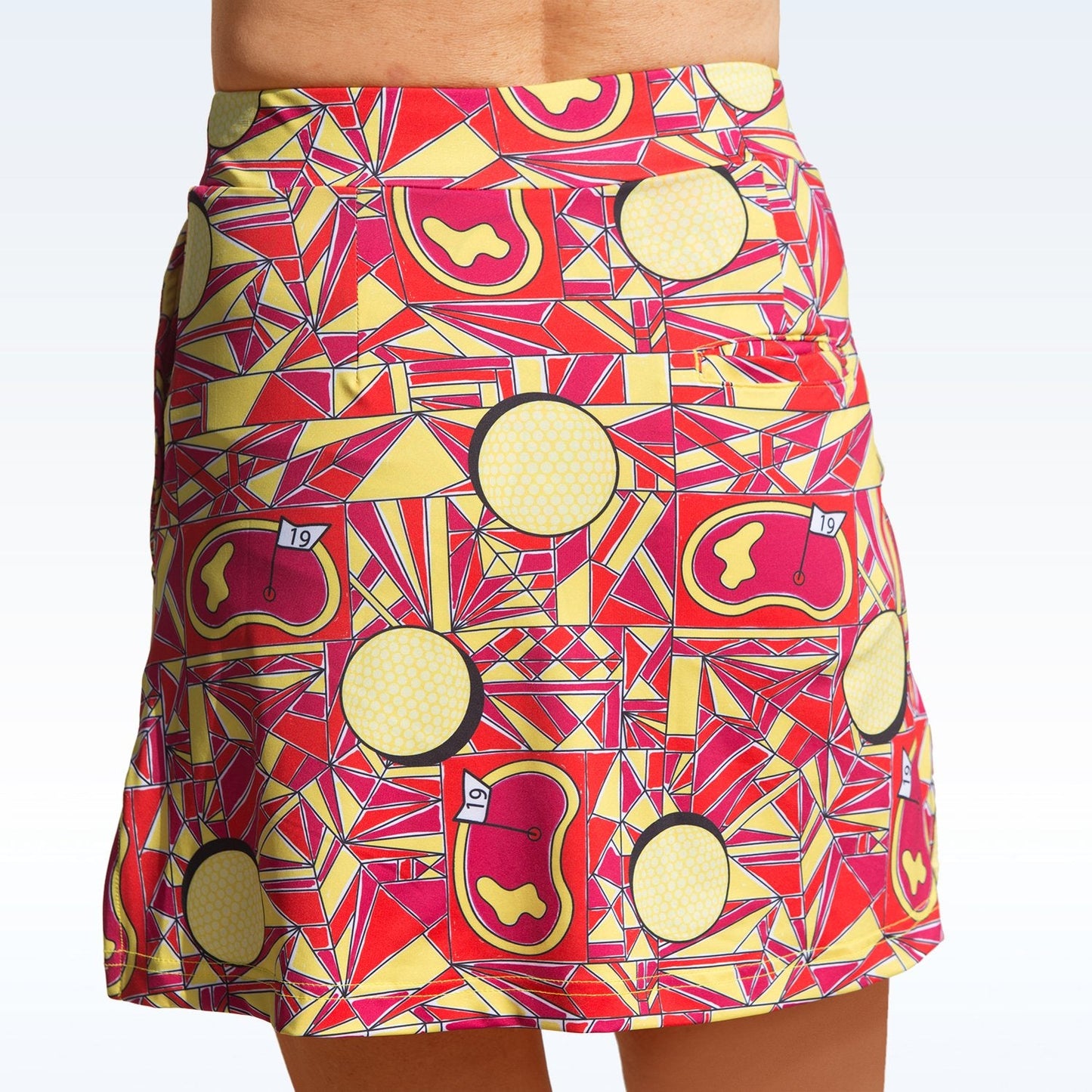 A person wears the Pickleball Bella Angle Shot Straight Golf Skort, featuring a figure-flattering fit and playful geometric patterns in red, yellow, and orange with yellow circles and "19" flag graphics for a sporty look.