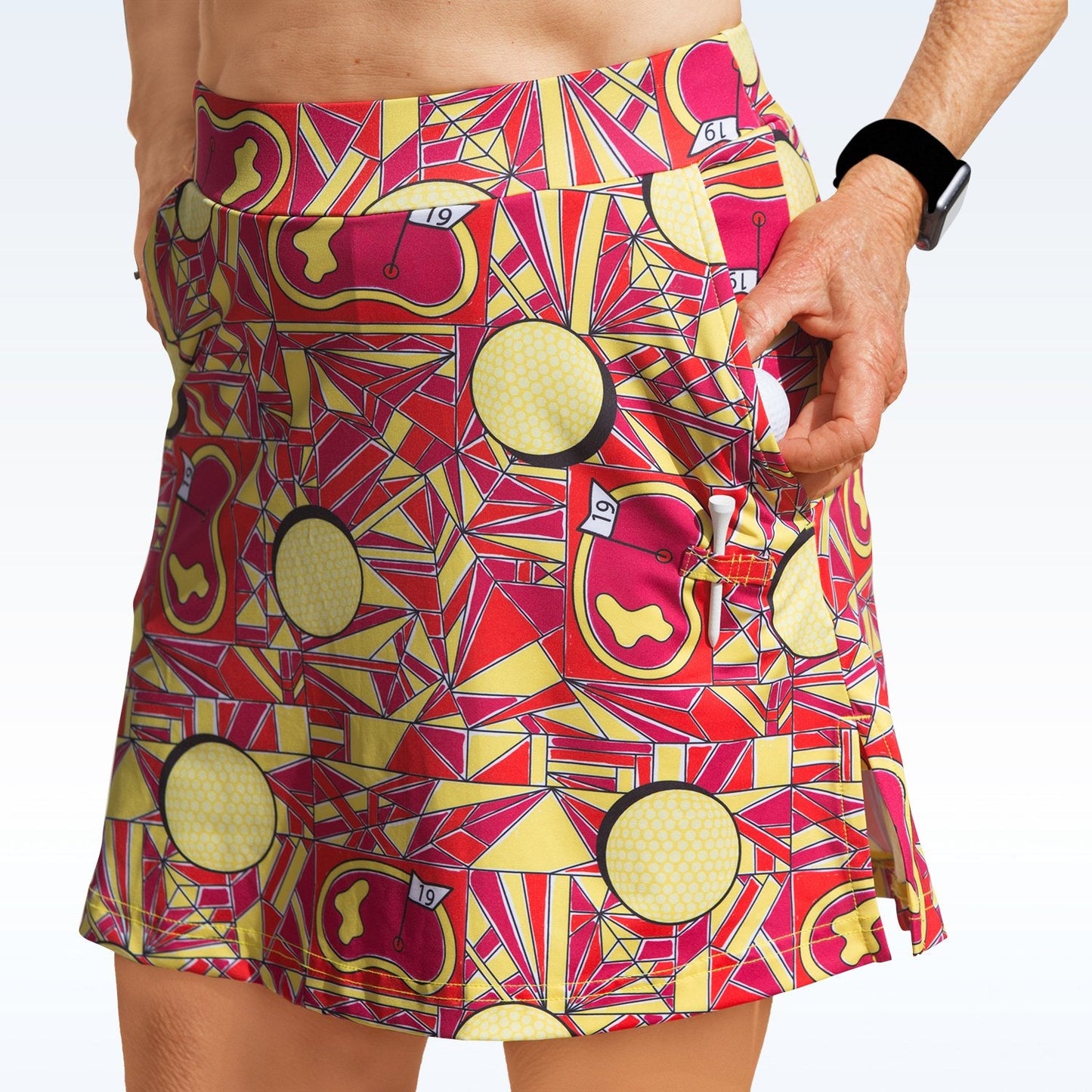 A person wears the Pickleball Bella Angle Shot Straight Golf Skort featuring red, yellow, and orange golf hole patterns. Made by Pickleball Bella from lightweight stretch fabric, it has pockets and is paired with a black smartwatch against a light background.