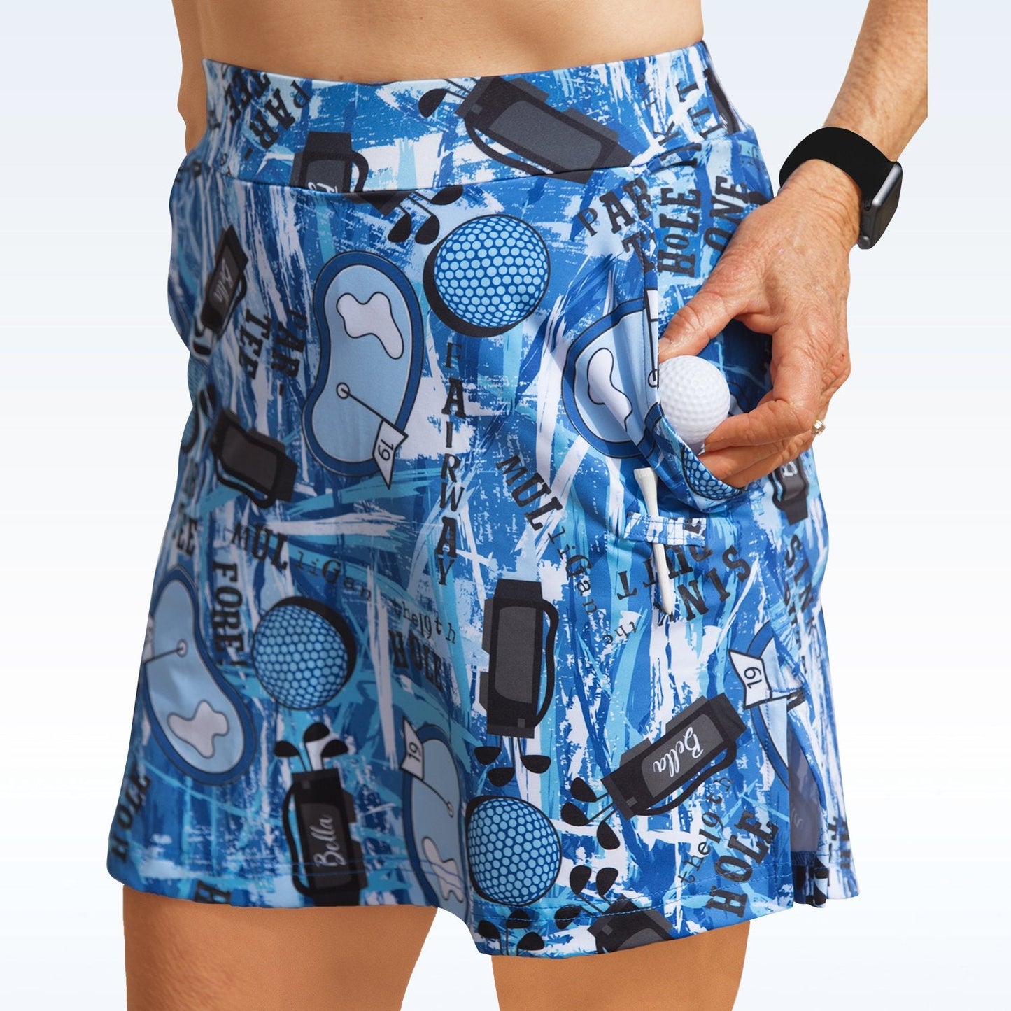 A person wears the Pickleball Bella Graffiti 2 Straight Golf Skort, featuring golf-themed graphics, and places a white golf ball into a pocket. Their midsection is visible and they're wearing a black wristwatch on their left wrist.
