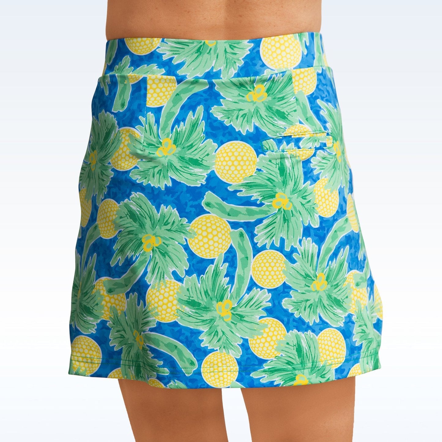 A person wears the Pickleball Bella Palms Straight Golf Skort by Pickleball Bella, featuring a vibrant pattern of yellow pineapples and green leaves. Only the skirt and lower torso are visible against a light gradient background.