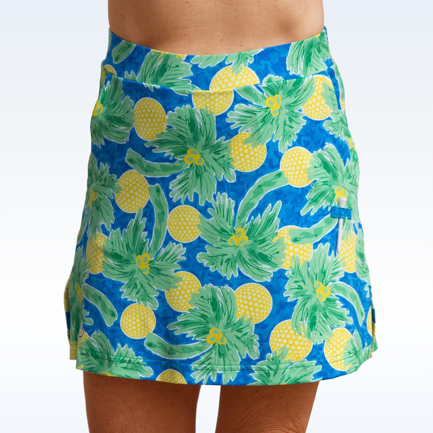 A person wears the Pickleball Bella Palms Straight Golf Skort by Pickleball Bella, featuring a blue base with yellow pineapples and green palm leaves; only the lower torso and upper legs are shown against a white to pale blue gradient background.