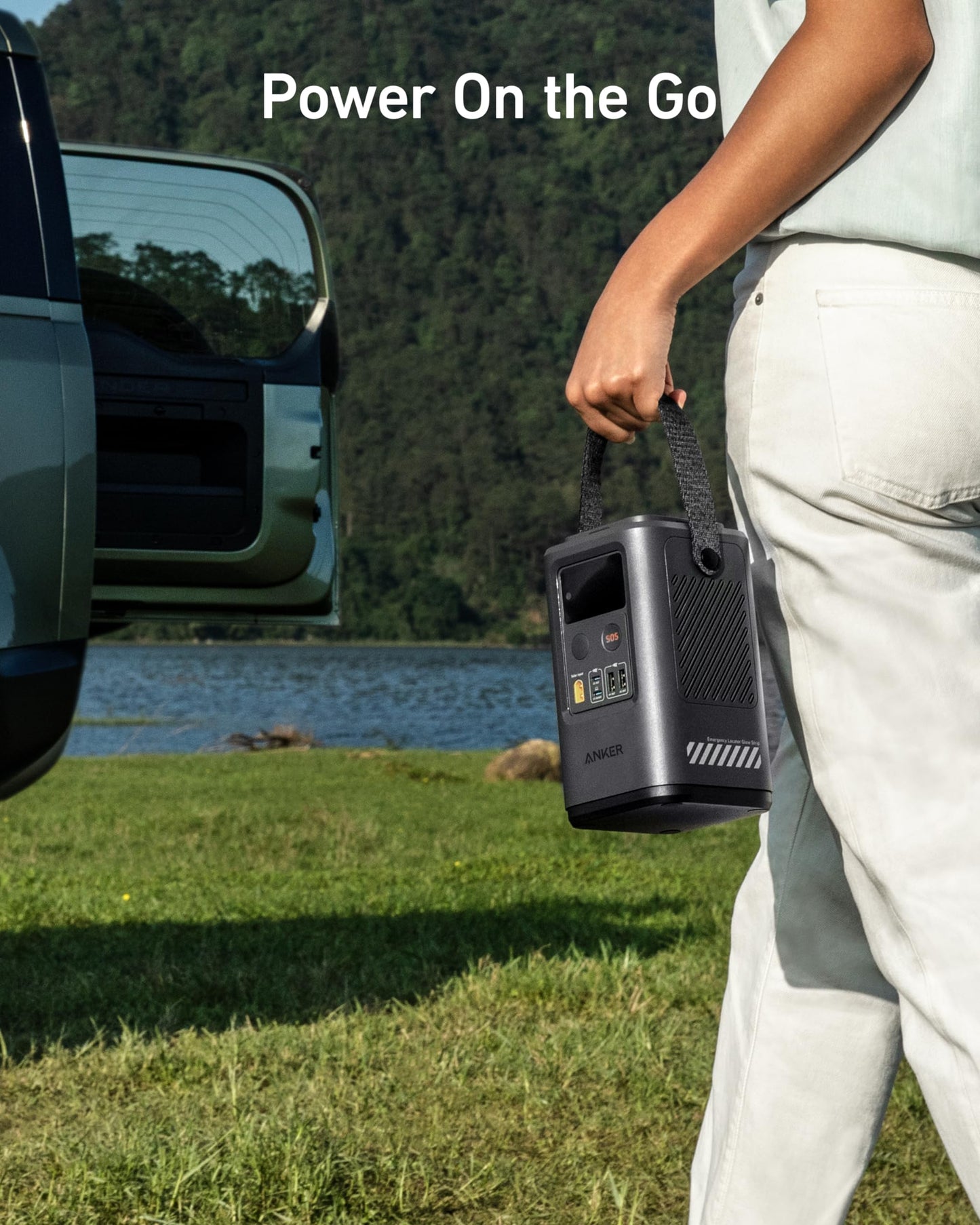 Wearing light clothes, a person carries the Anker 548 Power Bank (PowerCore Reserve 192Wh) with a strap, walking by a parked car near a lake and green hills. Text: “Power On the Go” highlights solar-charged portable energy for outdoor adventures.