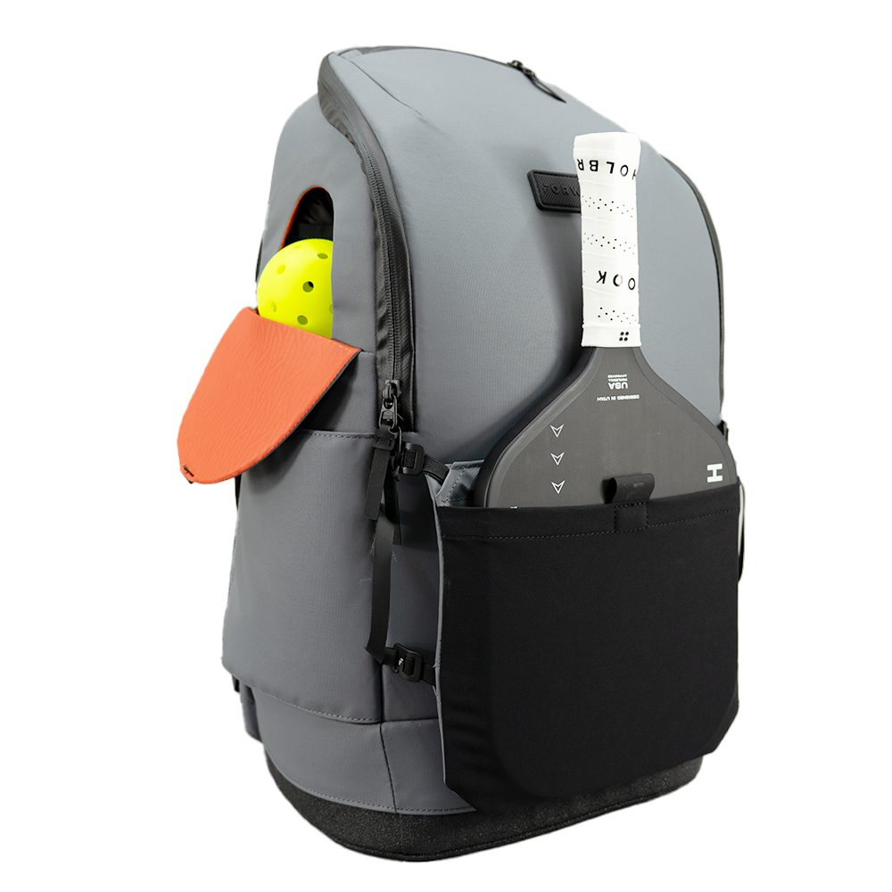 The FORWRD Court Caddy Pickleball Bag stands upright, featuring a modular gray design with black accents. An orange paddle and yellow pickleball are stored in the side pocket; the black front pocket has a white tag on the top zipper.