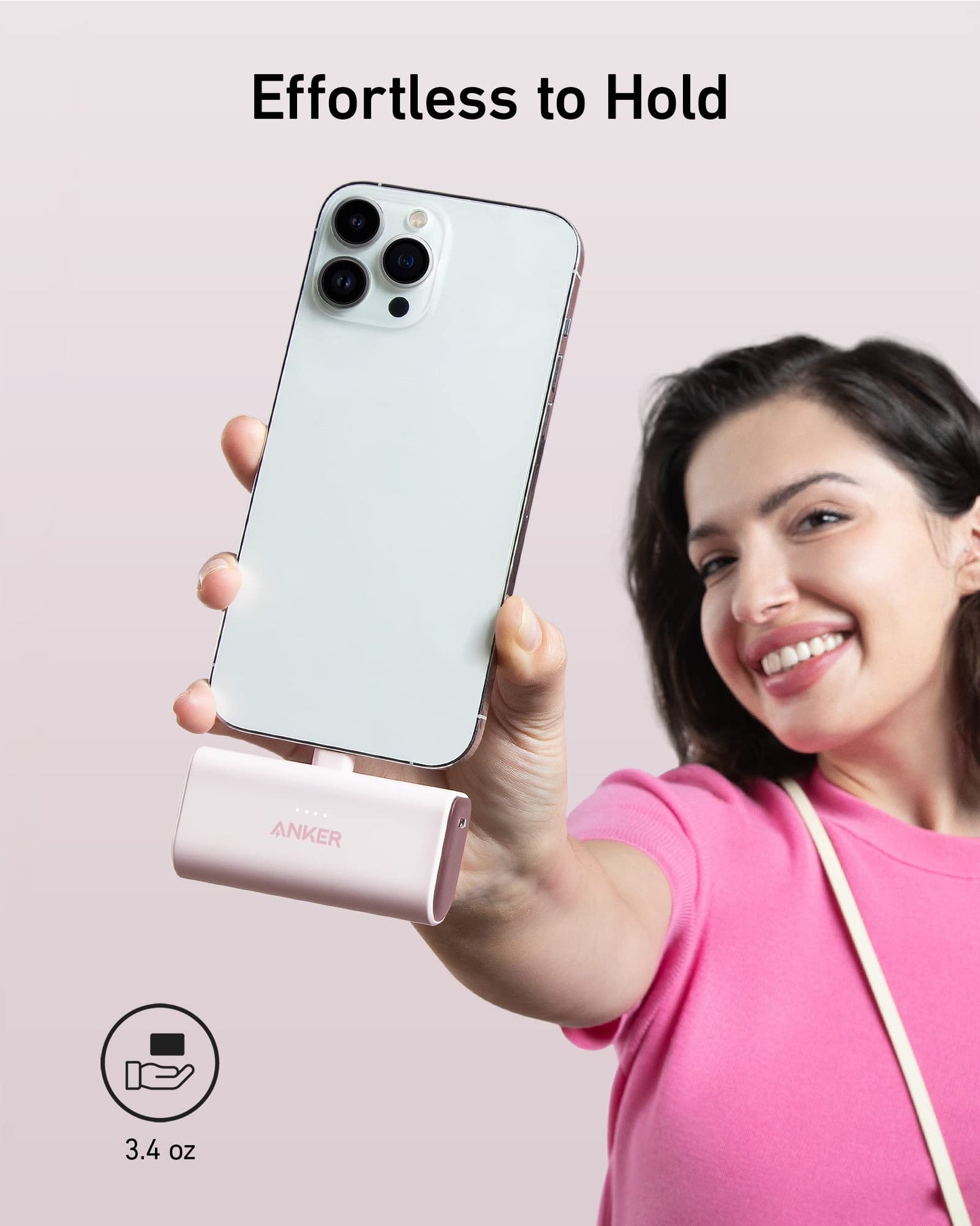 A smiling woman in a pink shirt holds an Anker Nano Power Bank (12W, Built-In Lightning Connector) attached to her white smartphone. The pale pink background reads "Effortless to Hold" with a 3.4 oz icon. Brand: Anker.