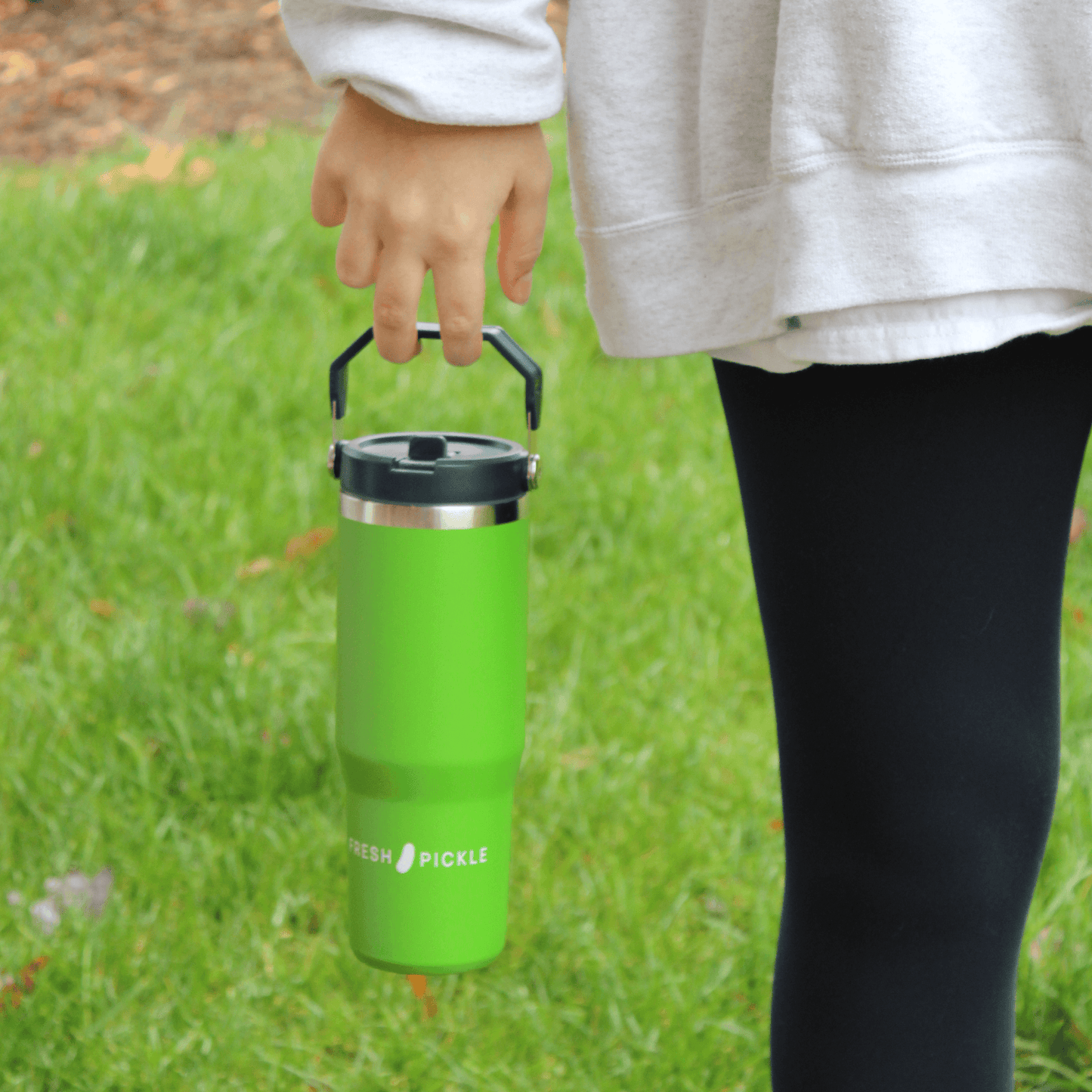 A person in a gray sweatshirt and black leggings holds the Fresh Pickle Tumbler by its black handle. The bright green 30 oz tumbler from Fresh Pickle is shown outdoors on grass with some orange and brown leaves nearby.