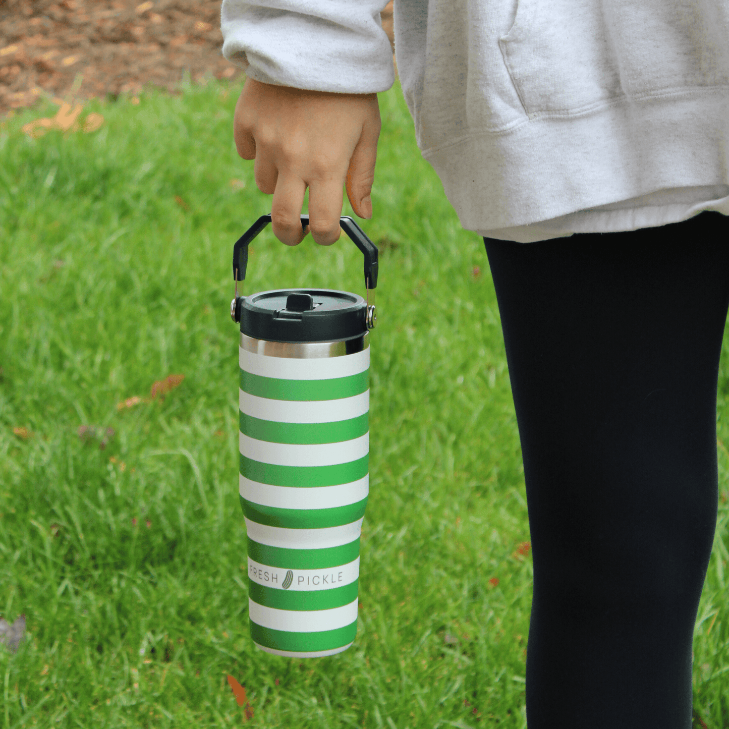 A person in black leggings and a gray hoodie holds the Fresh Pickle Tumbler by Fresh Pickle, a 30 oz bottle with a flip-up straw, while standing on a grassy lawn with scattered leaves. Only their hand, lower arm, and part of their body are visible.