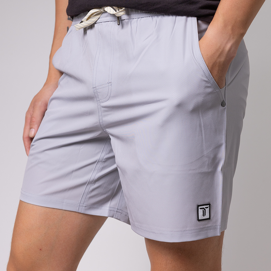 A person wears light gray Thrive Elite Shorts (7 in.) by Thrive Pickleball, featuring quick-drying stretch fabric, side pockets, a drawstring waistband, and a black-and-white logo near the hem. Their hands are in the pockets against a neutral background.