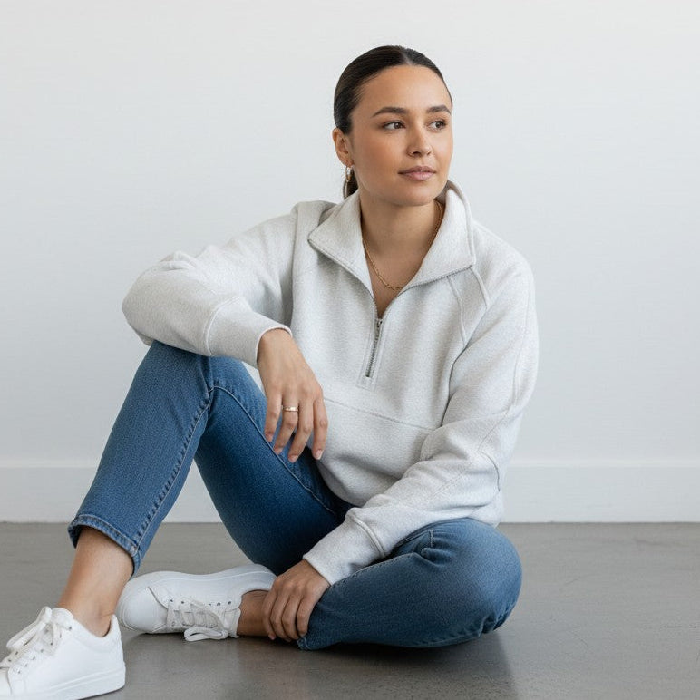 A woman with dark hair tied back sits against a light gray wall, wearing the DYM Athletics Cozy Up Full Length 1/2 Zip Sweatshirt in light gray, blue jeans, and white sneakers. She smiles softly and rests one arm on her knee.