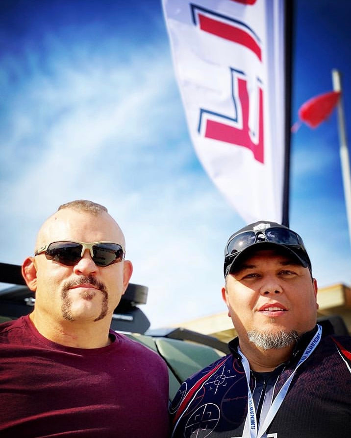 Two men pose outdoors under a blue sky. One wears a maroon shirt and Epoch Eyewear Grunt Tactical Sport Sunglasses; the other sports dark sunglasses, a black cap, and camo shirt. A large white flag waves behind them.