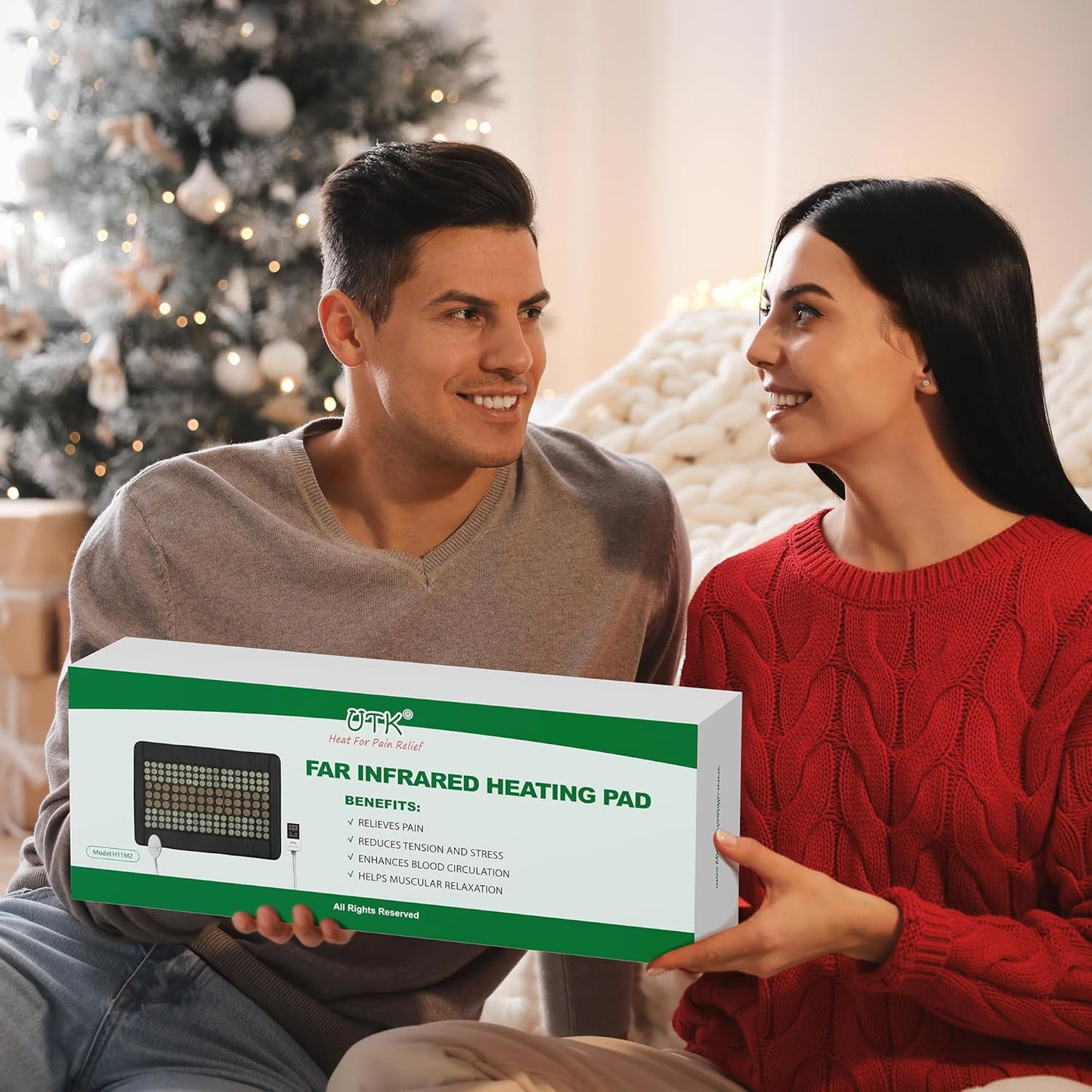 A smiling man and woman sit in a festive living room by a Christmas tree, holding the iReliev Far Infrared Natural Jade and Tourmaline Heating Pad (31" x 21"), highlighting soothing muscle pain relief for the holidays.