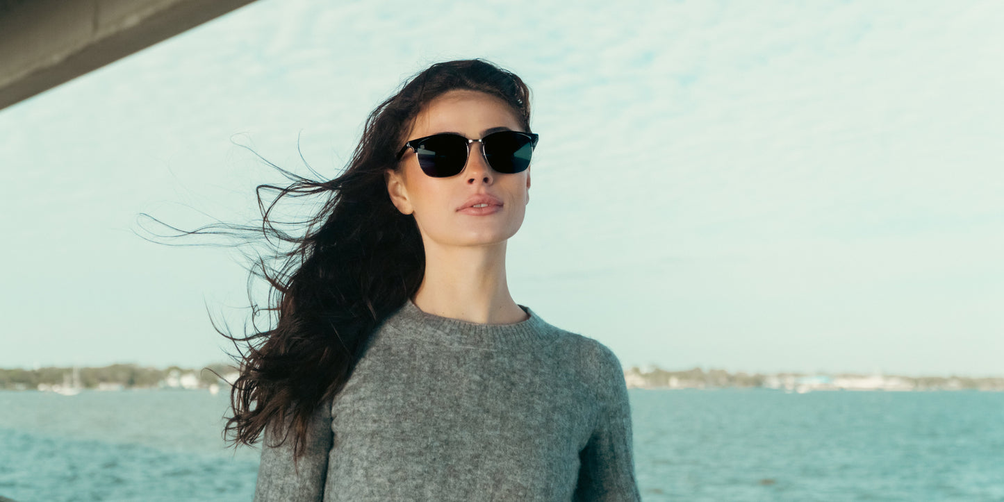 A woman with long dark hair, wearing Neven Eyewear Bullet black polarized sunglasses and a grey sweater, stands outdoors by the water as wind blows her hair and a cloudy sky reveals distant land and trees.