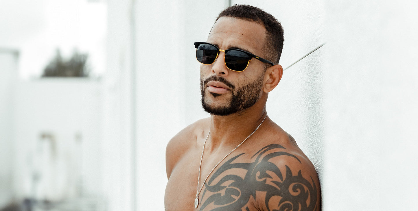 A shirtless, bearded man with short hair and a large black tribal tattoo on his right shoulder models Neven Eyewear's Gold Rush sunglasses—stylish polarized UV400 TR90 frames—plus a silver necklace, set against a minimal white background.