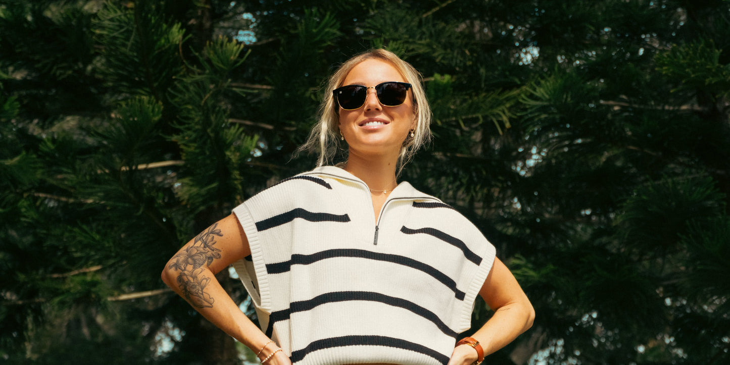 A smiling person with tattoos and blonde hair pulled back stands outdoors in sunlight, wearing Neven Eyewear's Gold Rush TR90 sunglasses with polarized UV400 protection and a white short-sleeved sweater, surrounded by lush green trees.