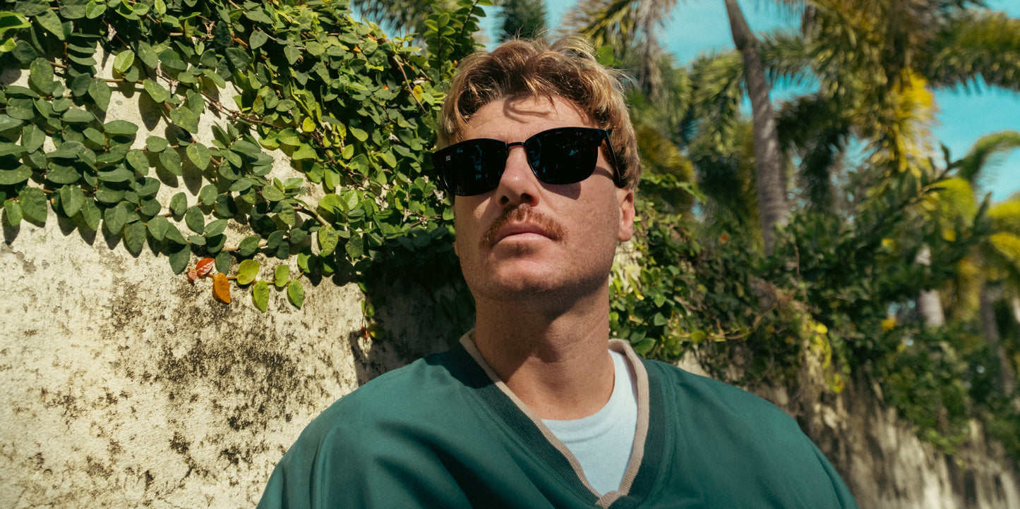 A man with light hair and a mustache wears Neven Eyewear Stealth black polarized sunglasses with UV400 protection and a green jacket over a white shirt, standing outdoors by a vine-covered stone wall and palm trees under a blue sky.