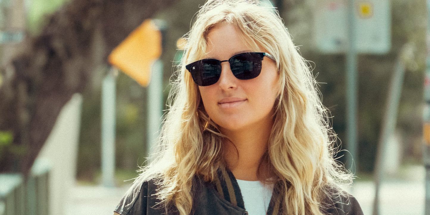 A person with wavy blonde hair wears Neven Eyewear Stealth black polarized sunglasses and a dark jacket outdoors. The background features green foliage and urban elements, softly blurred in bright natural light.