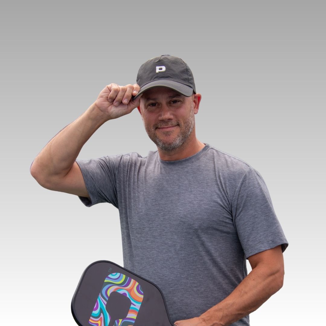 A man in a gray moisture-wicking tee and the Playly Store Hi Hat Pickleball Hat smiles, holding the hat's brim. In his other hand, he holds a colorful pickleball paddle, set against a smooth gray-to-white gradient background.