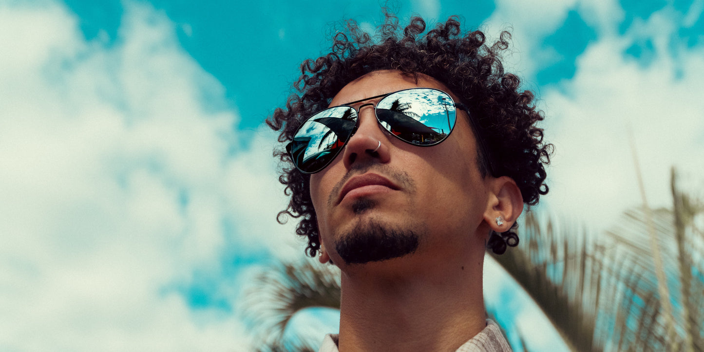 A man with curly hair, a goatee, and a nose piercing wears Neven Eyewear Mavs sunglasses with stainless steel frames. Blue sky and palm tree reflect in his polarized UV400 lenses as he looks upward.