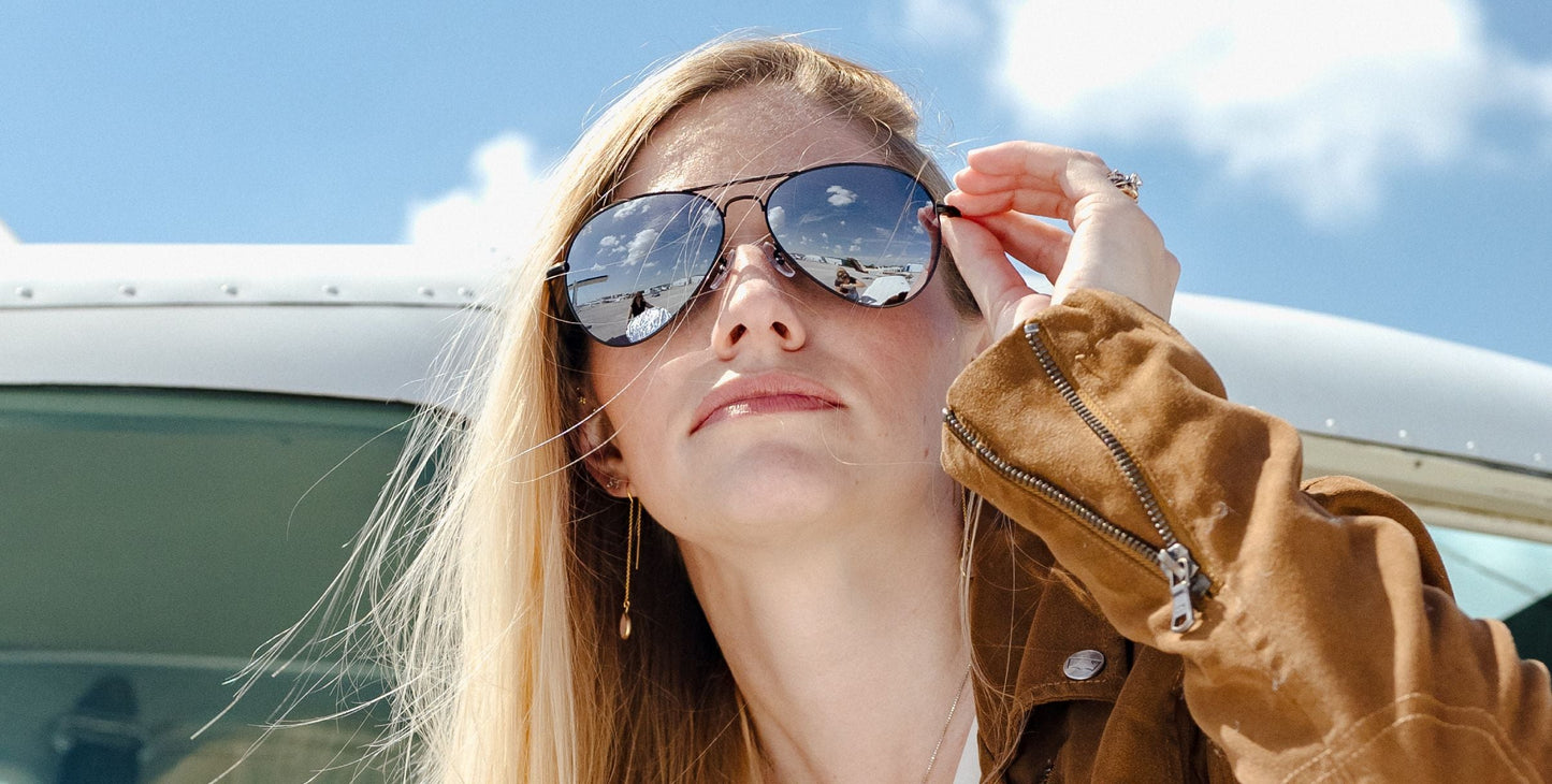 A woman with long blonde hair in a brown jacket stands by a small airplane, touching her Neven Eyewear Mavs sunglasses with polarized UV400 lenses, which reflect the blue sky and clouds as she gazes upward.