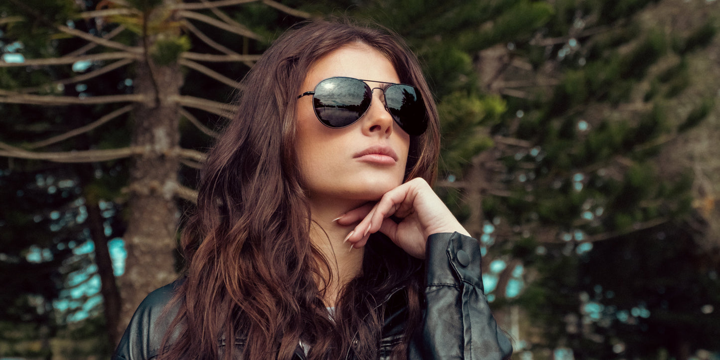 A woman with long wavy brown hair wears Neven Eyewear’s Nightfall polarized sunglasses with UV400 protection and a black leather jacket, gazing thoughtfully into the distance amidst tall evergreens.