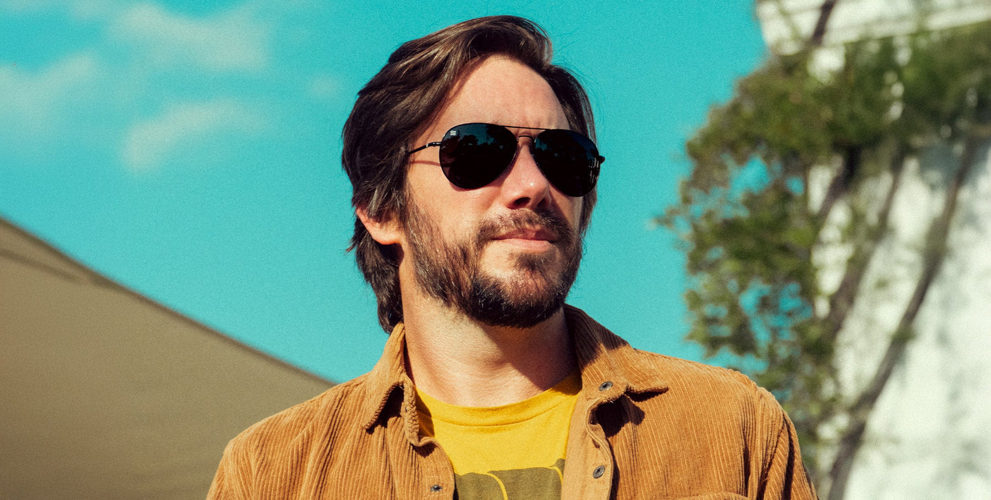 A bearded man wears Neven Eyewear's Nightfall polarized sunglasses, a yellow shirt, and a brown corduroy jacket while standing outdoors on a sunny day, with blue sky and blurred trees in the background.