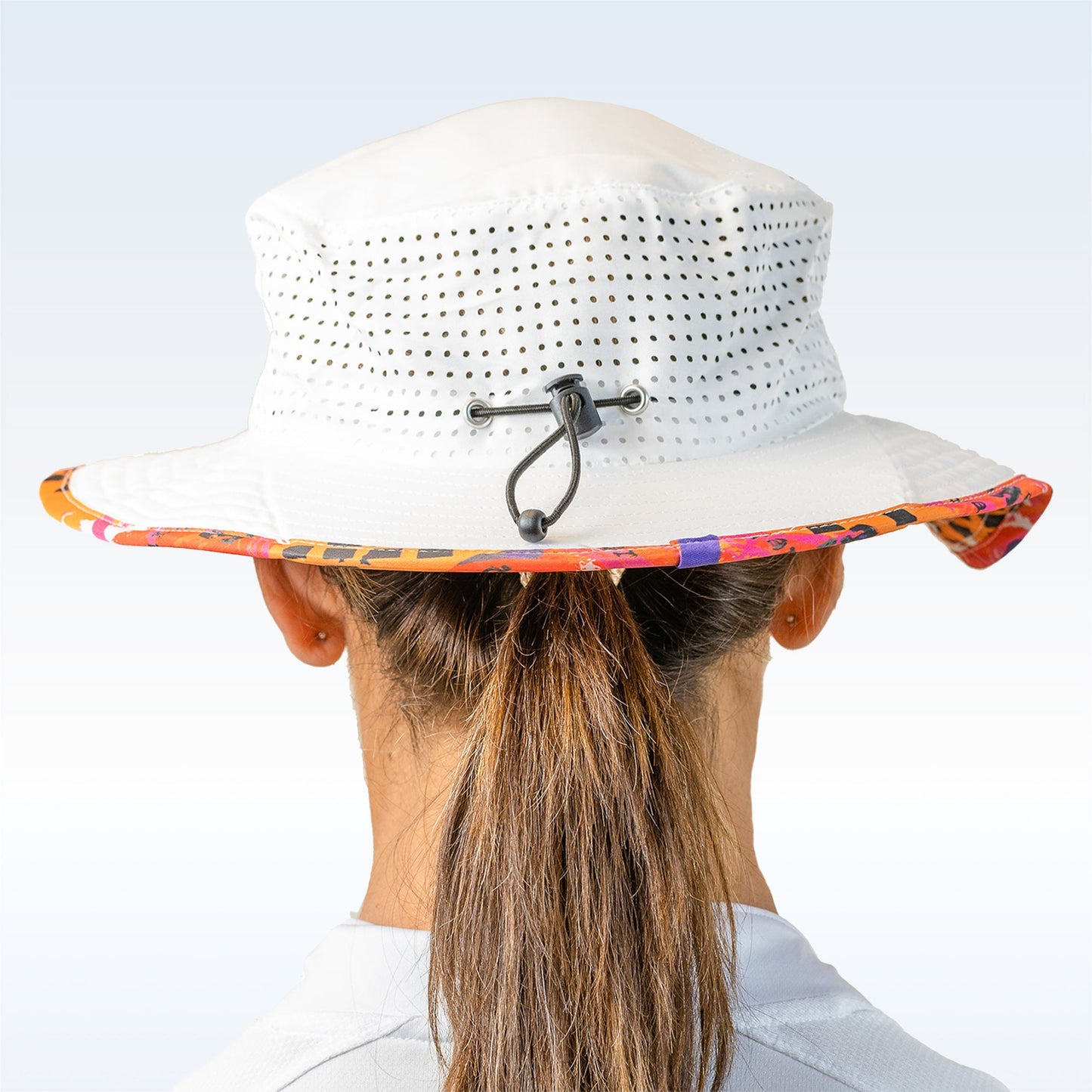 A person with brown hair in a ponytail wears the Pickleball Bella PB Bucket Hat Graffiti 3, a white perforated bucket hat with a colorful patterned brim and an adjustable drawstring, paired with a light-colored shirt, viewed from the back.