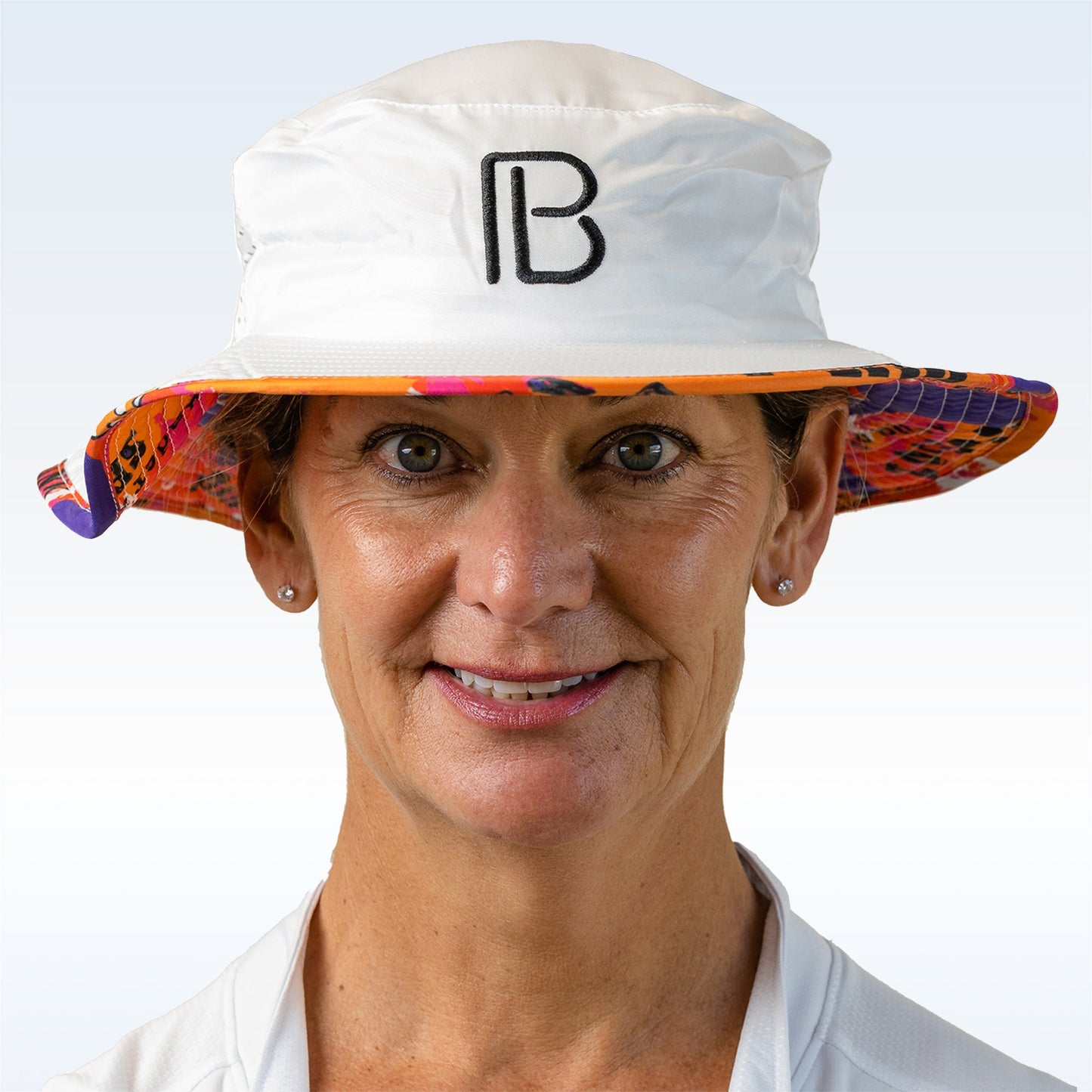 A middle-aged woman with short hair smiles at the camera, wearing the Pickleball Bella PB Bucket Hat Graffiti 3, featuring sun protection, a black "B" logo, and a colorful patterned brim. She pairs it with a white collared shirt and stud earrings.