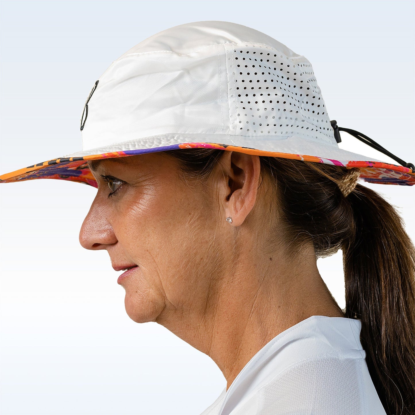A middle-aged woman, in profile, wears the Pickleball Bella PB Bucket Hat Graffiti 3 featuring a white crown, multicolored brim, perforated side panels, and an adjustable drawstring for sun protection. Her brown hair is in a low ponytail with a white shirt.