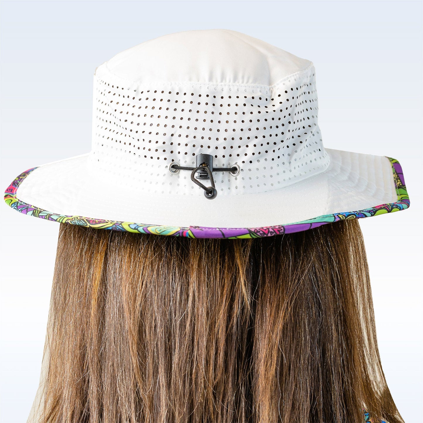 A person with long brown hair is seen from behind wearing the Pickleball Bella PB Bucket Hat Groovy, featuring ventilation holes, a black back clip for sun protection, a floral-trimmed brim, and a white design against a plain light blue background.