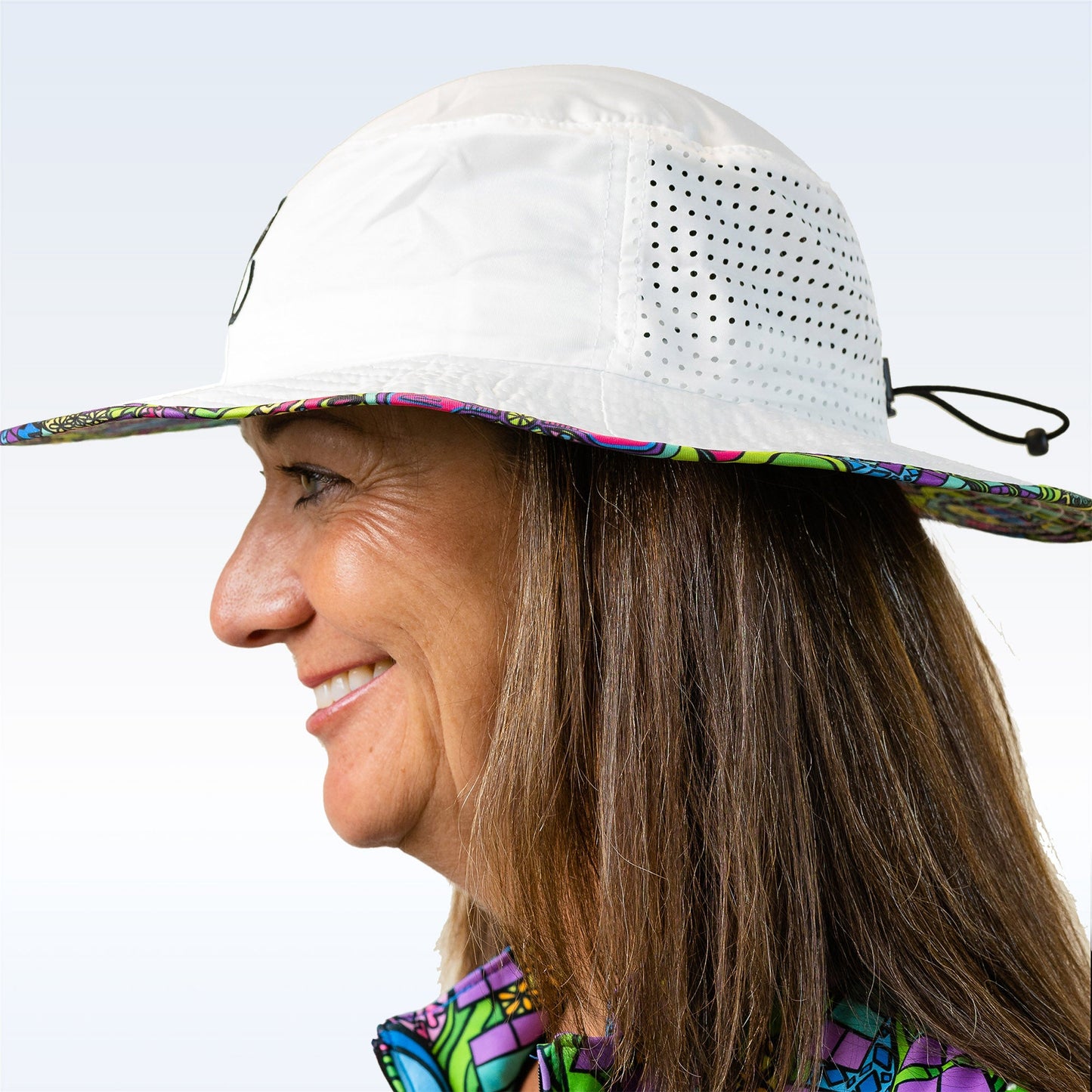 A smiling woman in profile sports the Pickleball Bella PB Bucket Hat Groovy, featuring ventilation holes, a drawstring for sun protection, and a colorful patterned brim. She has straight brown hair and wears a vibrant top—ideal for pickleball.