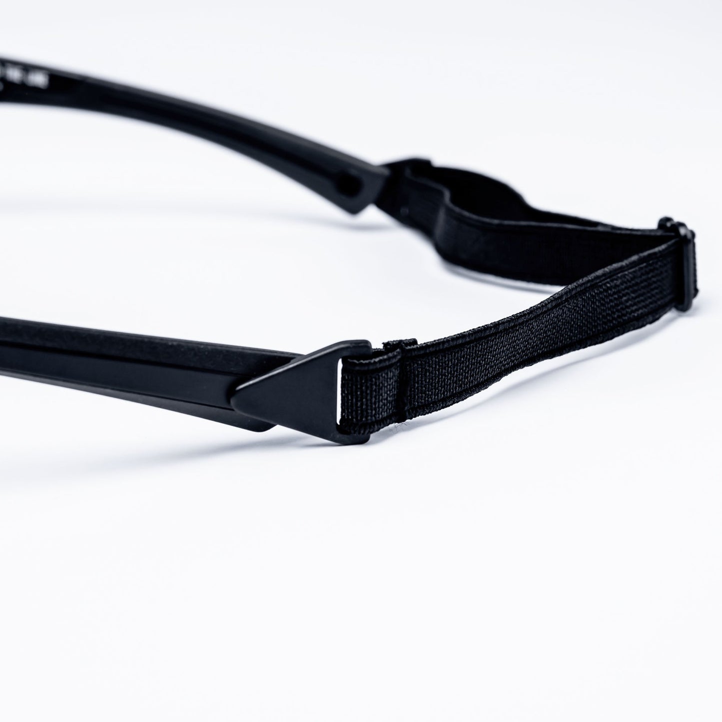 A close-up photo shows a black BRIOTI FOCOS Head Strap with a plastic buckle attached to the arm of safety or sports glasses on a white background, highlighting the adjustable strap and its connection point.
