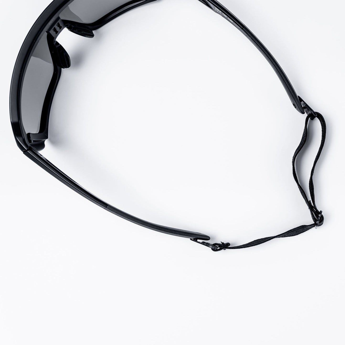 A close-up shows the BRIOTI FOCOS Head Strap attached to black eyewear with dark lenses on a white surface. The image focuses on the right, highlighting the thin adjustable strap and nose pads, showing part of the front and right arm.