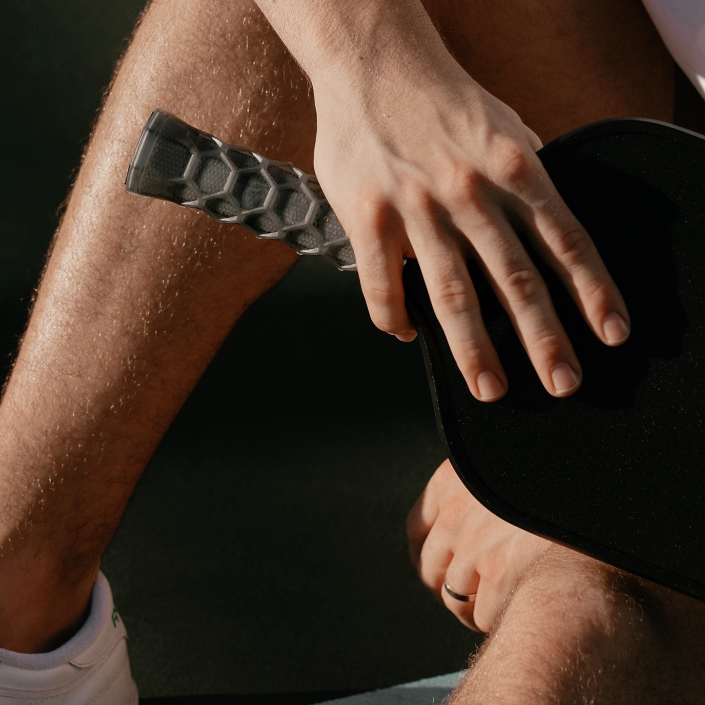 A close-up of a hand resting on the Hesacore Grip Pickleball Carbon Grip (5.25” long, stiff feel, charcoal), with an ergonomic handle visible in warm light. The person’s bare leg, wrist, and part of a white sneaker are also shown.