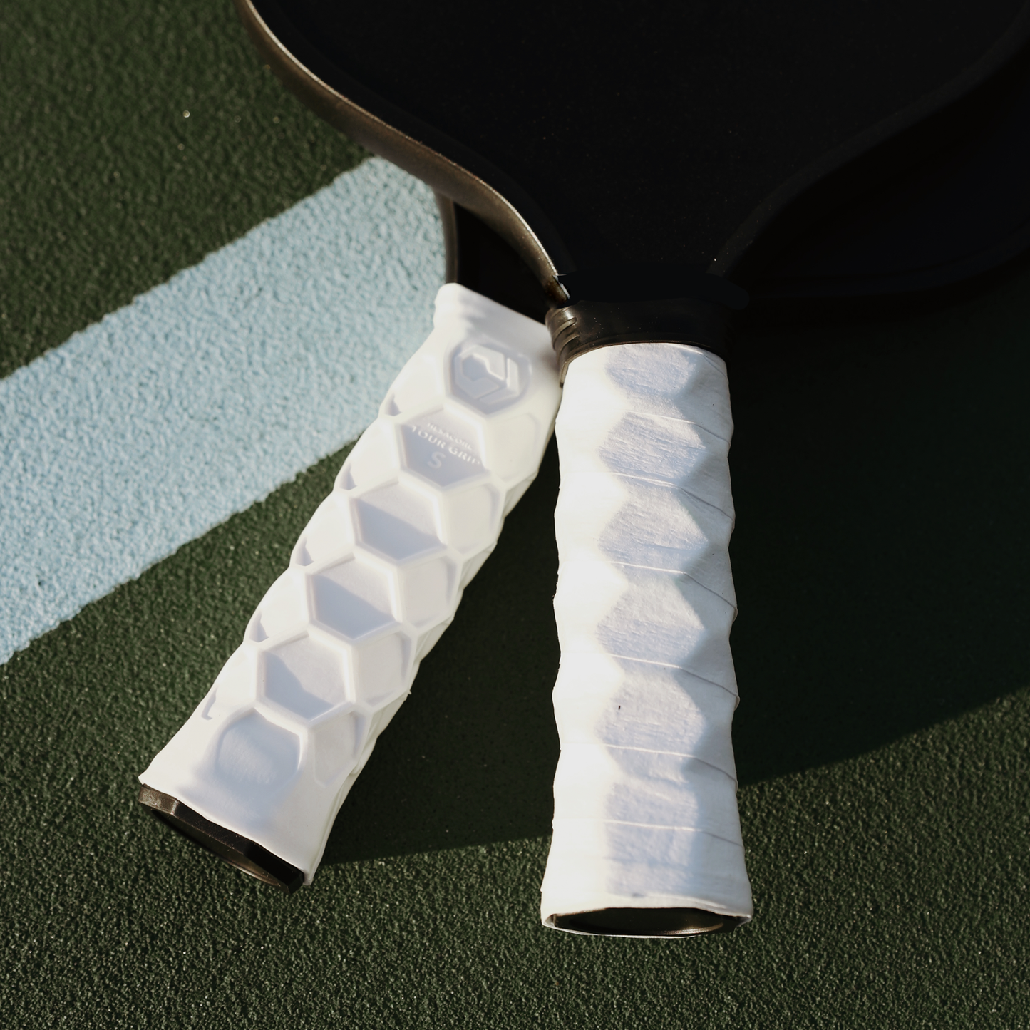 Two black pickleball paddles with white Hesacore Grip 5.25-inch Regular Feel grips rest on a green court, partially overlapping over a diagonal light blue line. Sunlight highlights the ergonomic, hexagonal-patterned grip details.