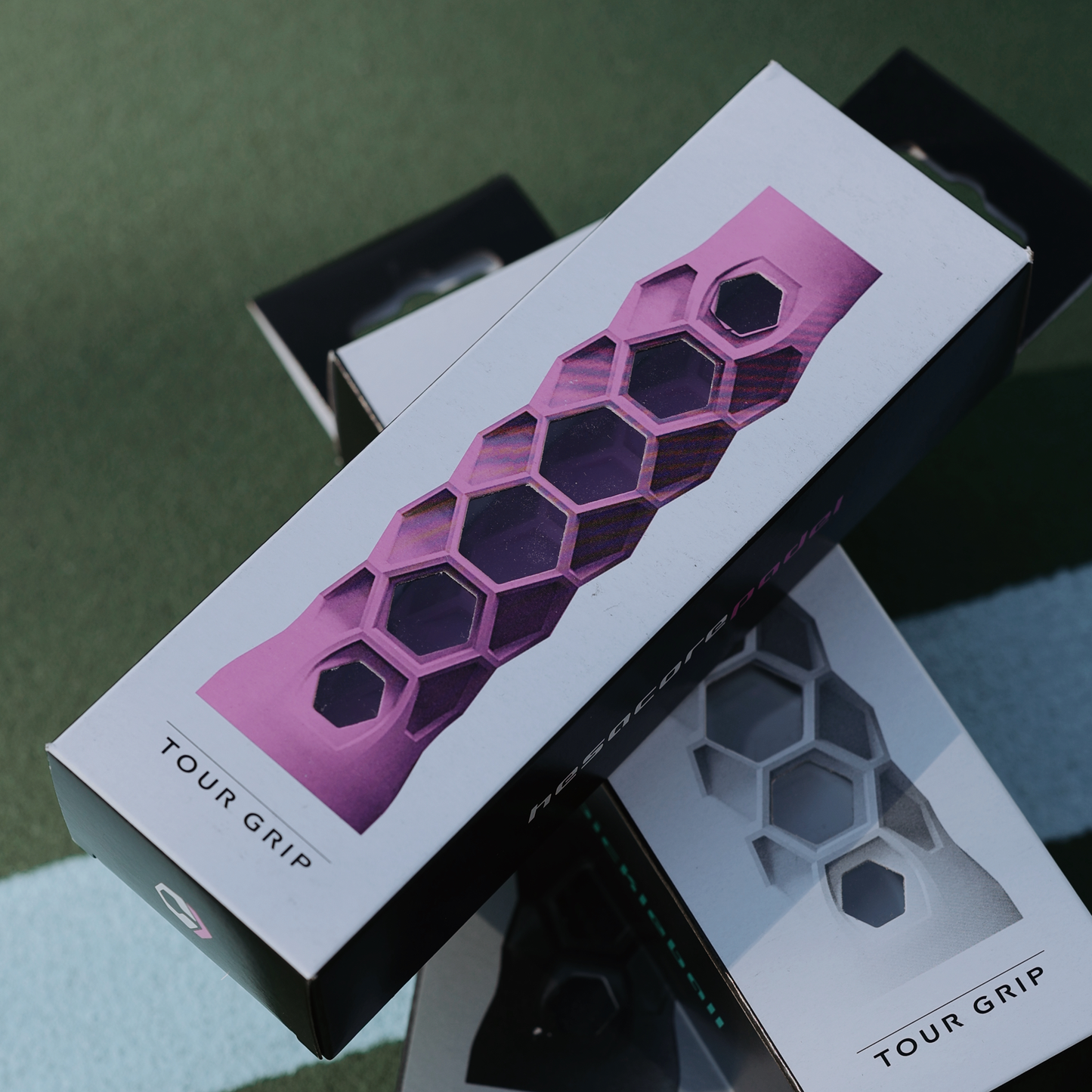 A rectangular box shows the Hesacore Grip Pickleball Grip (5.25" Long, Regular Feel, Pink) with a hexagonal, sweat-absorbing TOUR GRIP against a tennis court. Nearby is another box with a gray ergonomic paddle grip.