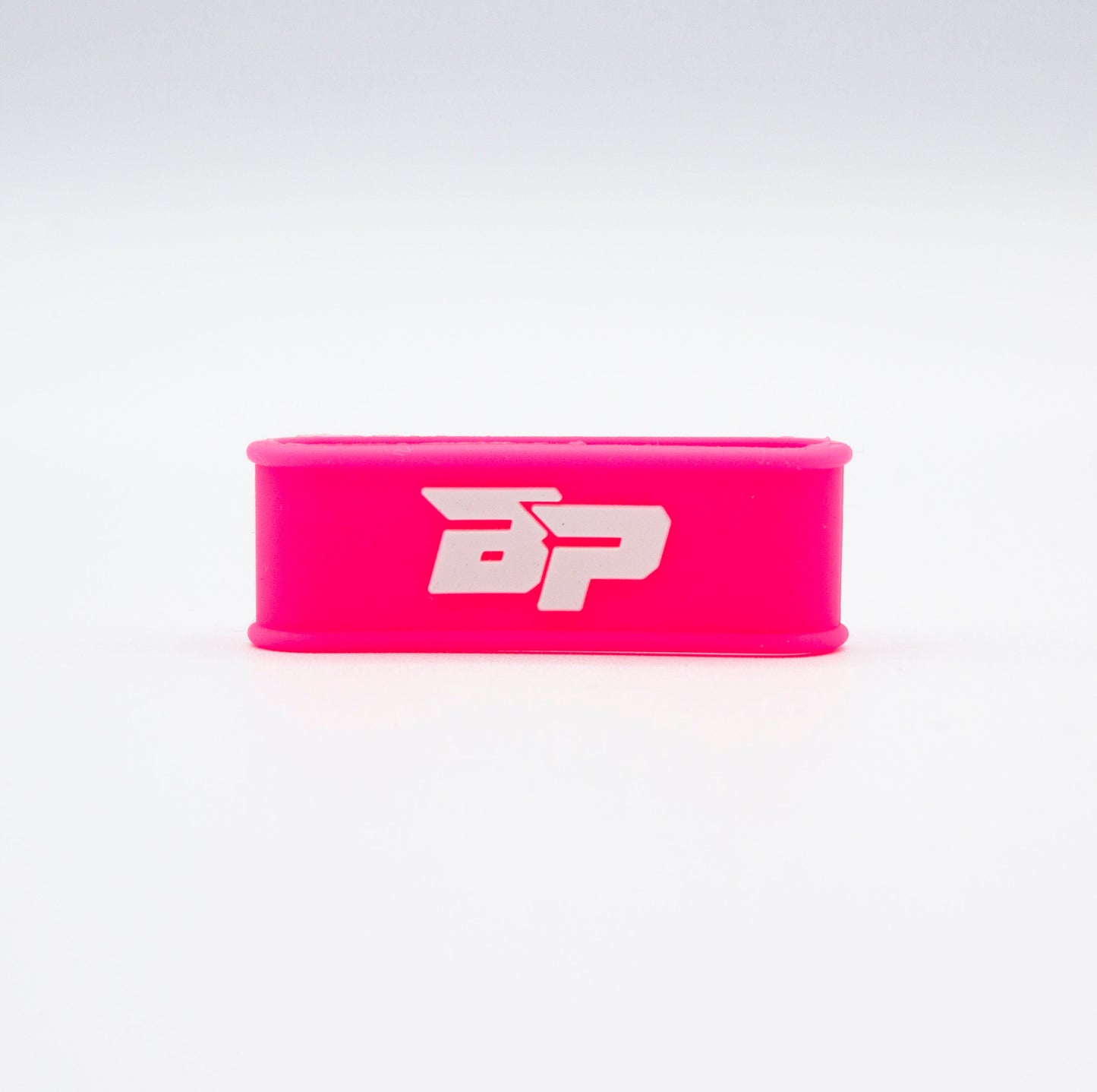 A bright pink, rectangular Paddle Band by Bodhi Performance stands upright on a white background. Featuring bold white “BP” lettering, this silicone grip band is a stylish, minimalistic pickleball paddle accessory with striking color contrast.