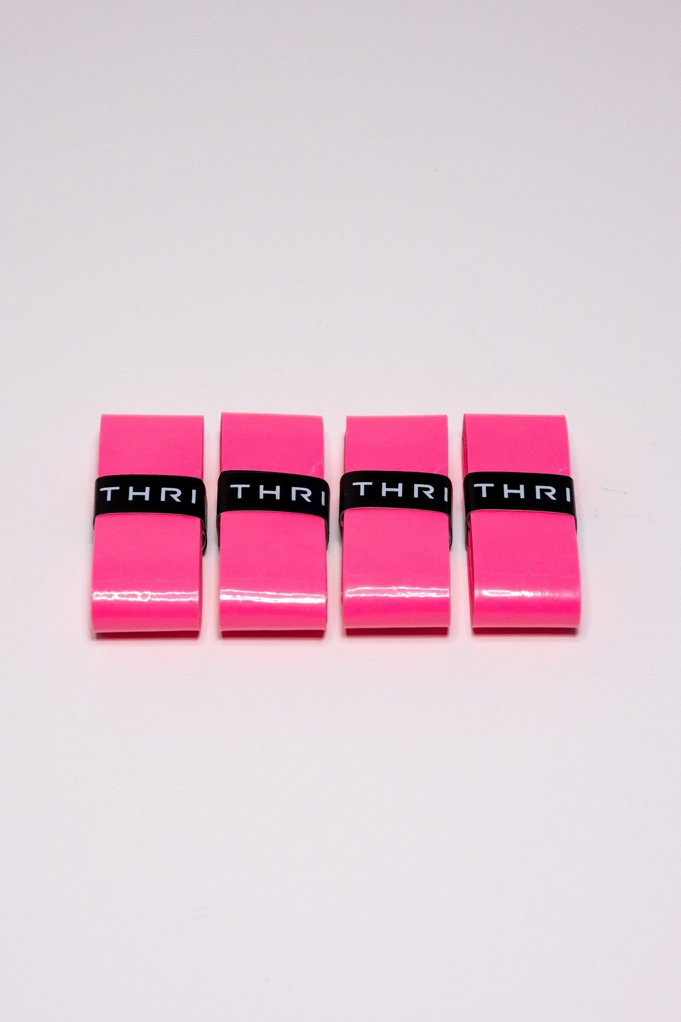 Four bright pink rectangular Thrive Pickleball Overgrips (Super Tacky) are lined up on a white surface. Each has a black band with "THRI" in white. The background is plain and uncluttered.
