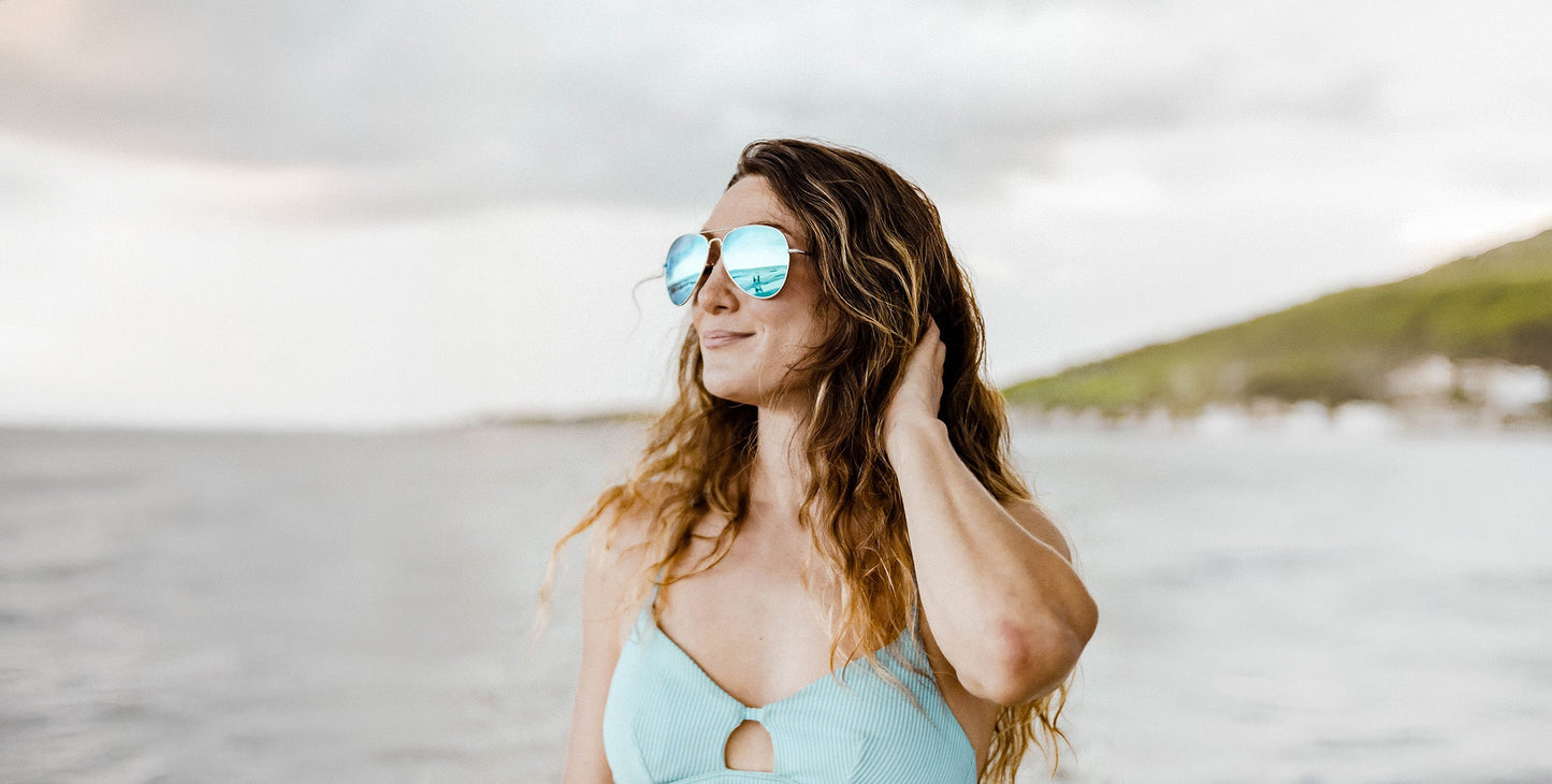 Wearing Neven Eyewear H2O polarized sunglasses, a woman with long wavy hair in a light blue swimsuit stands by the water, touching her hair. Behind her, the blurred ocean and a green hillside are visible under a cloudy sky.
