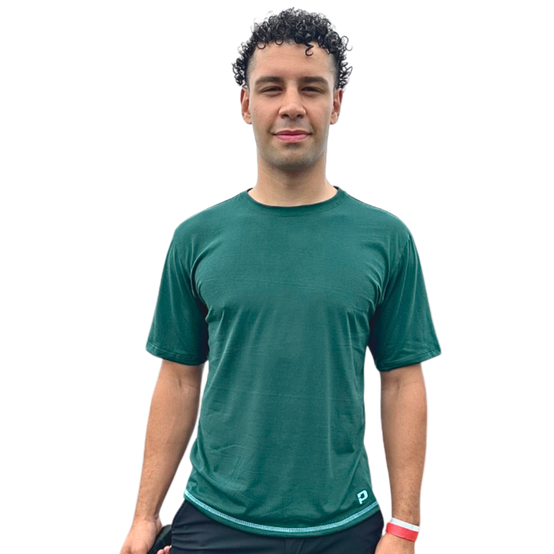 A man with short, curly dark hair faces forward wearing the Playly Store Warrior Tee 2.0 in green and black shorts, paired with a red wristband on his left wrist, posed against a smooth gray gradient background. His expression is neutral.