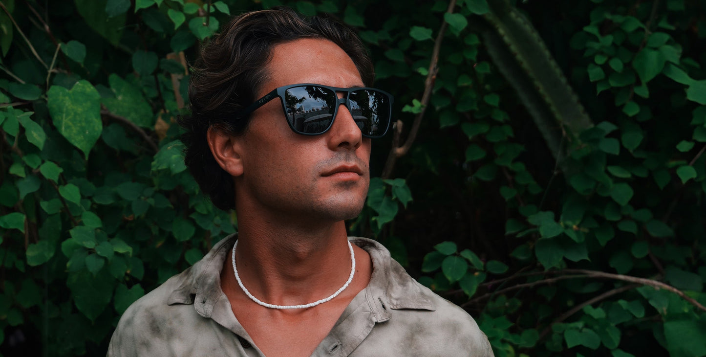 A man with wavy brown hair wears Neven Eyewear's Abyss all-black polarized UV400 sunglasses, a white beaded necklace, and a grey patterned shirt, standing before lush green foliage. Reflections of trees and sky appear in his shades.