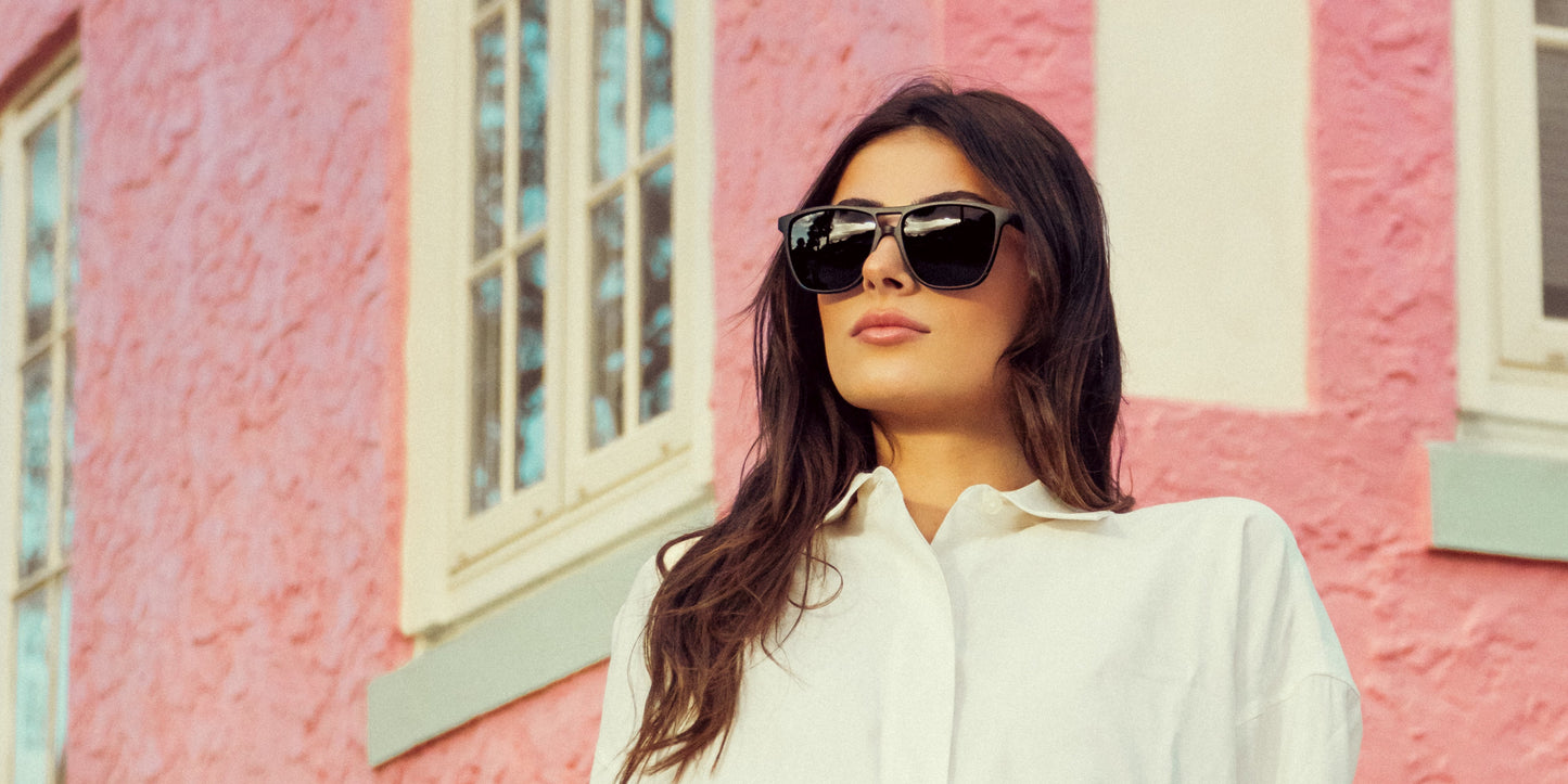 A woman with long dark hair and a white button-up shirt stands in front of a pink building, wearing Neven Eyewear's Abyss oversized all-black sunglasses with polarized UV400 protection for a sunny, stylish vibe.