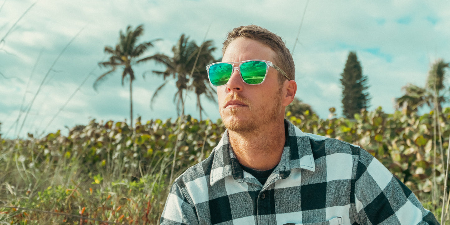 A man in a black-and-white plaid shirt wears Neven Eyewear Hangar's sunglasses with emerald green lenses that reflect a grassy field, adding color to his relaxed look as he sits outdoors among palm trees and clouds.