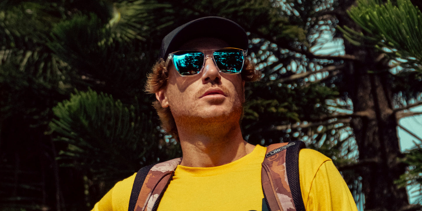 A man wearing Neven Eyewear’s Lucid mirrored rectangular sunglasses and a black cap stands outdoors, sunlight highlighting his confident expression as he looks upward in front of green trees.