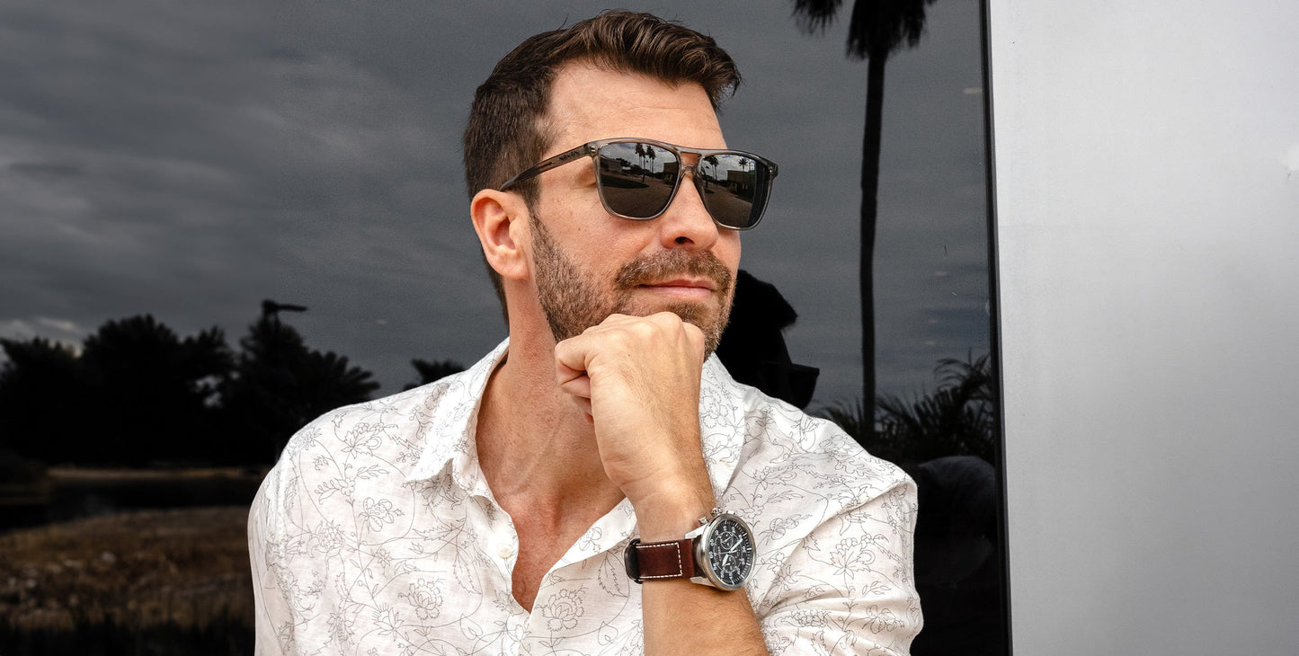 A man with short brown hair and a trimmed beard wears Neven Eyewear Rip Tide polarized sunglasses and a white patterned shirt, posing thoughtfully with a wristwatch. The background includes trees, cloudy sky, and a reflective surface.