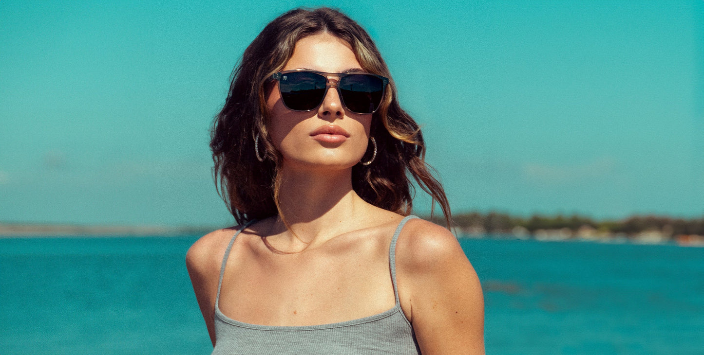 A woman with long brown hair stands outdoors in bright sunlight at Indian Harbor Beach, wearing Neven Eyewear Rip Tide polarized sunglasses and a gray sleeveless top, with large hoop earrings and a calm blue body of water behind her.