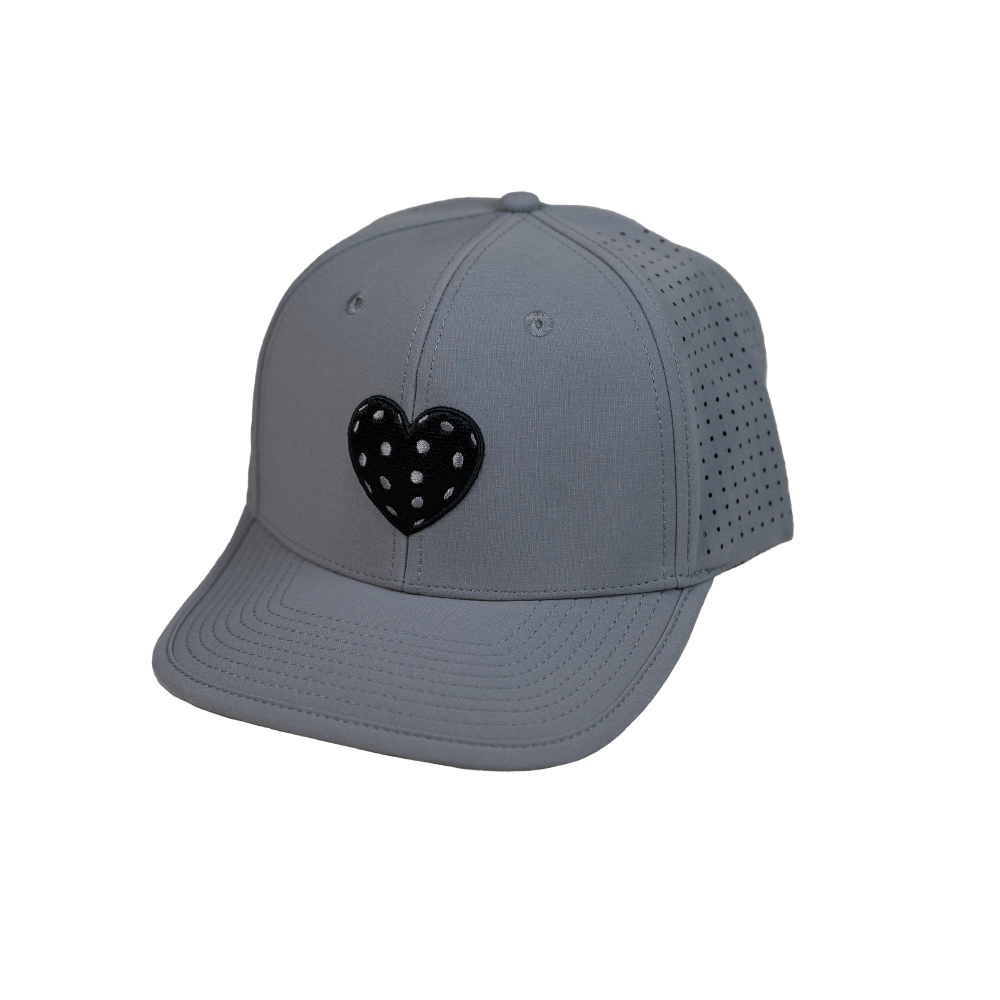 The SEXY Brand I ❤️ Pickleball AquaSnapBack in Gray is a light gray baseball cap with a curved brim and perforated sides. It features a glossy black heart with white polka dots above the brim, combining sporty style with modern flair.