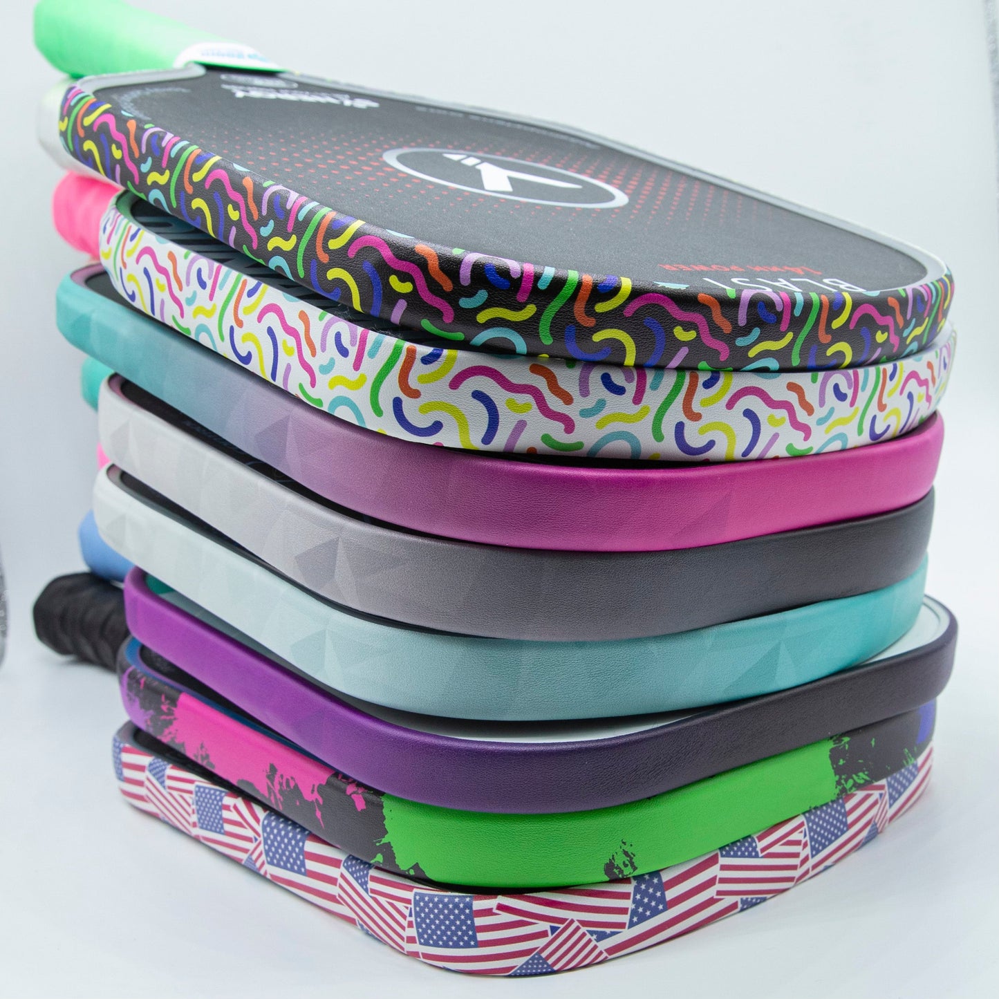 A stack of colorful pickleball paddles with abstract, geometric, and American flag designs are neatly fitted with Bodhi Performance Premium Edge Tape - PATTERNS for paddle protection, all arranged vertically on a white background.