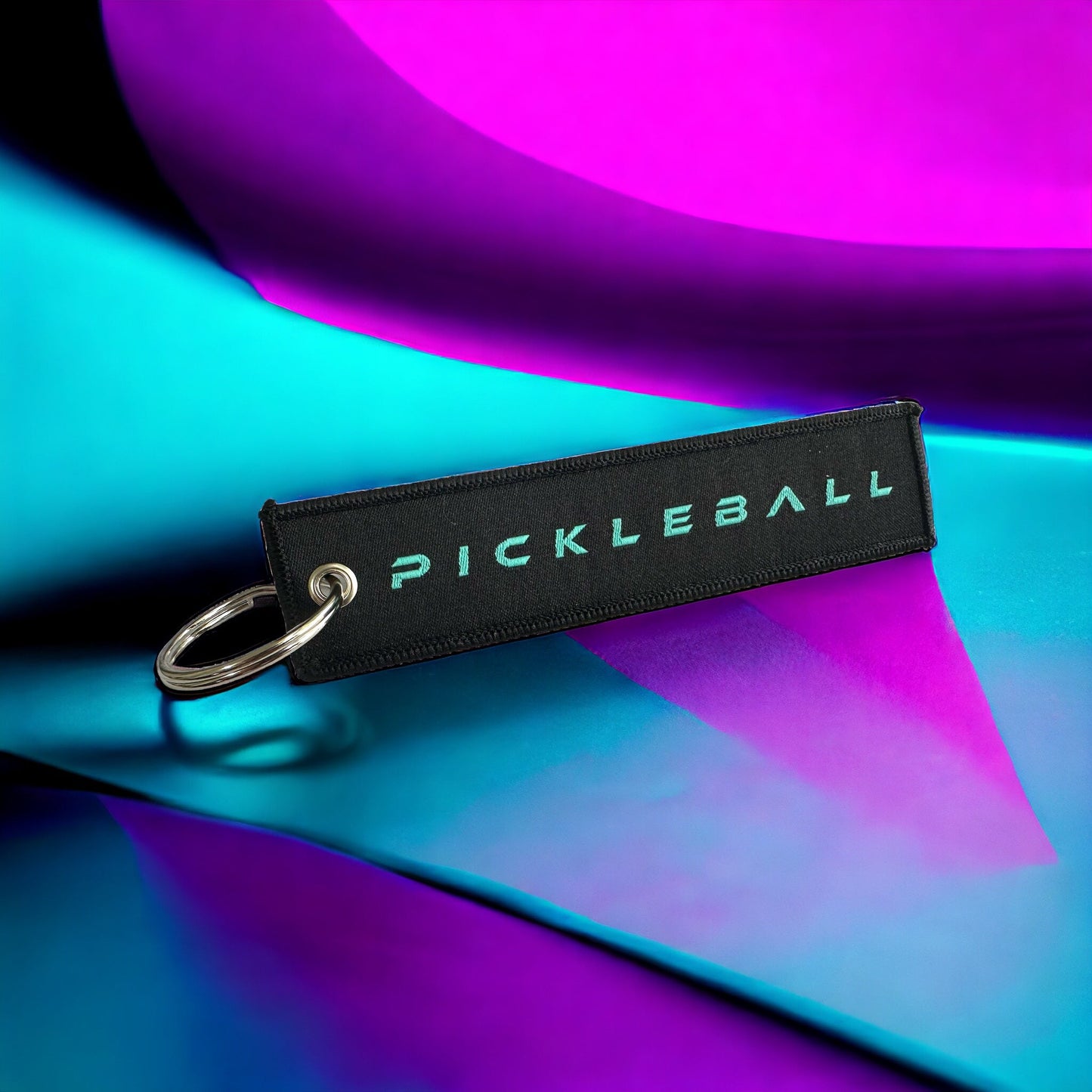 A Potenza Pickleball Logo Key Chain with bold white text on black fabric and a custom metal keyring rests on a reflective surface under vivid blue and purple lighting, highlighting smooth, abstract color gradients.