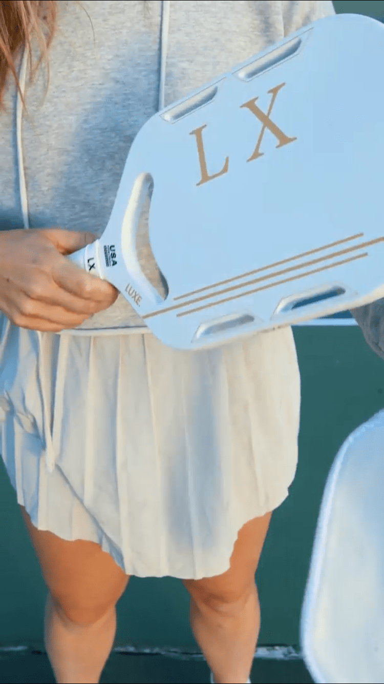 A woman in a gray hoodie and white pleated skirt holds the lightweight, USA Pickleball-approved LUXE Pickleball LX paddle with a gold “LX” logo on an outdoor court. Only her torso, hand, and part of her legs are visible. Her face is out of frame.