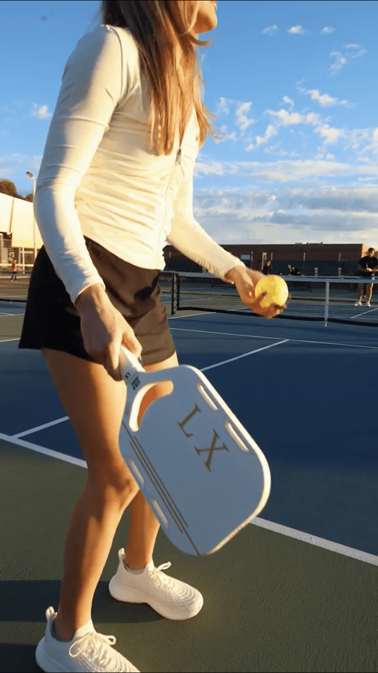 On a blue pickleball court, a woman in a white long-sleeve shirt, black shorts, and white shoes holds the LUXE Pickleball LX paddle (USA Pickleball approved) in her right hand and a yellow ball in her left. Another player stands by the net in the background.