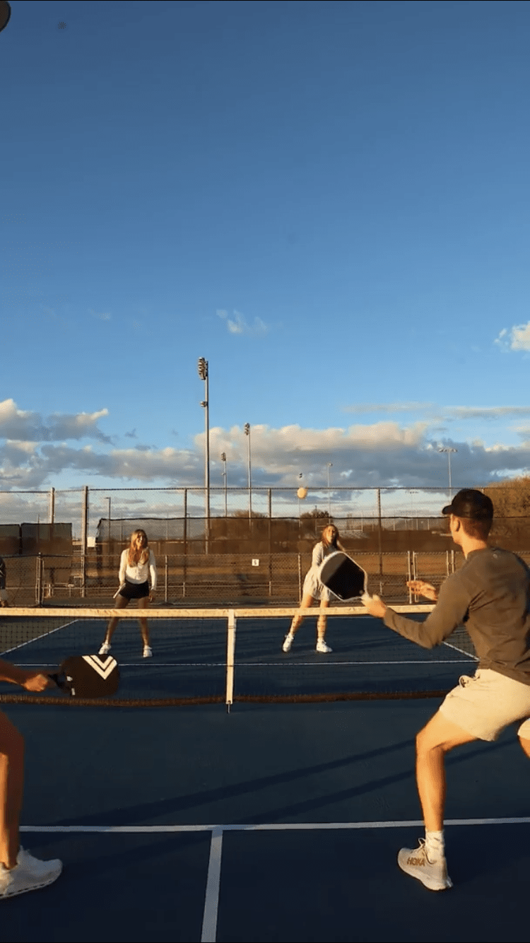 Four people play pickleball in pairs on an outdoor court at sunset. Two players hold LUXE Pickleball Dinker carbon fiber paddles as the ball soars mid-air, under blue sky and athletic lights. USA Pickleball Approved equipment in action.